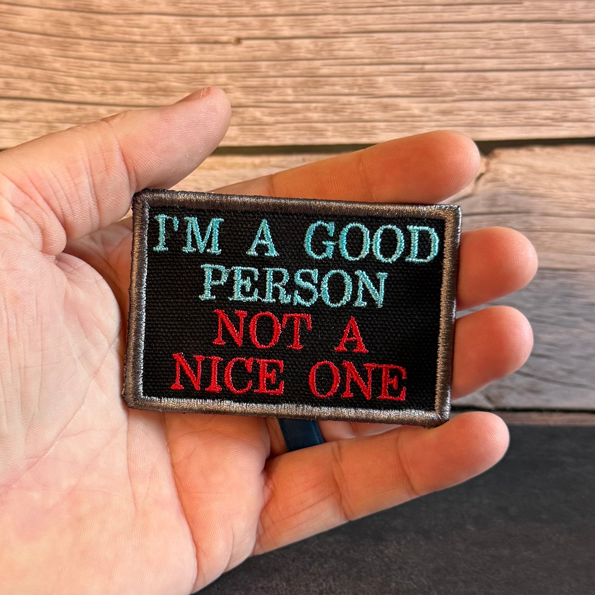 A hand holding a rectangular embroidered patch with the text 'I'M A GOOD PERSON NOT A NICE ONE' in light blue and red on a black background, set against a wooden surface.