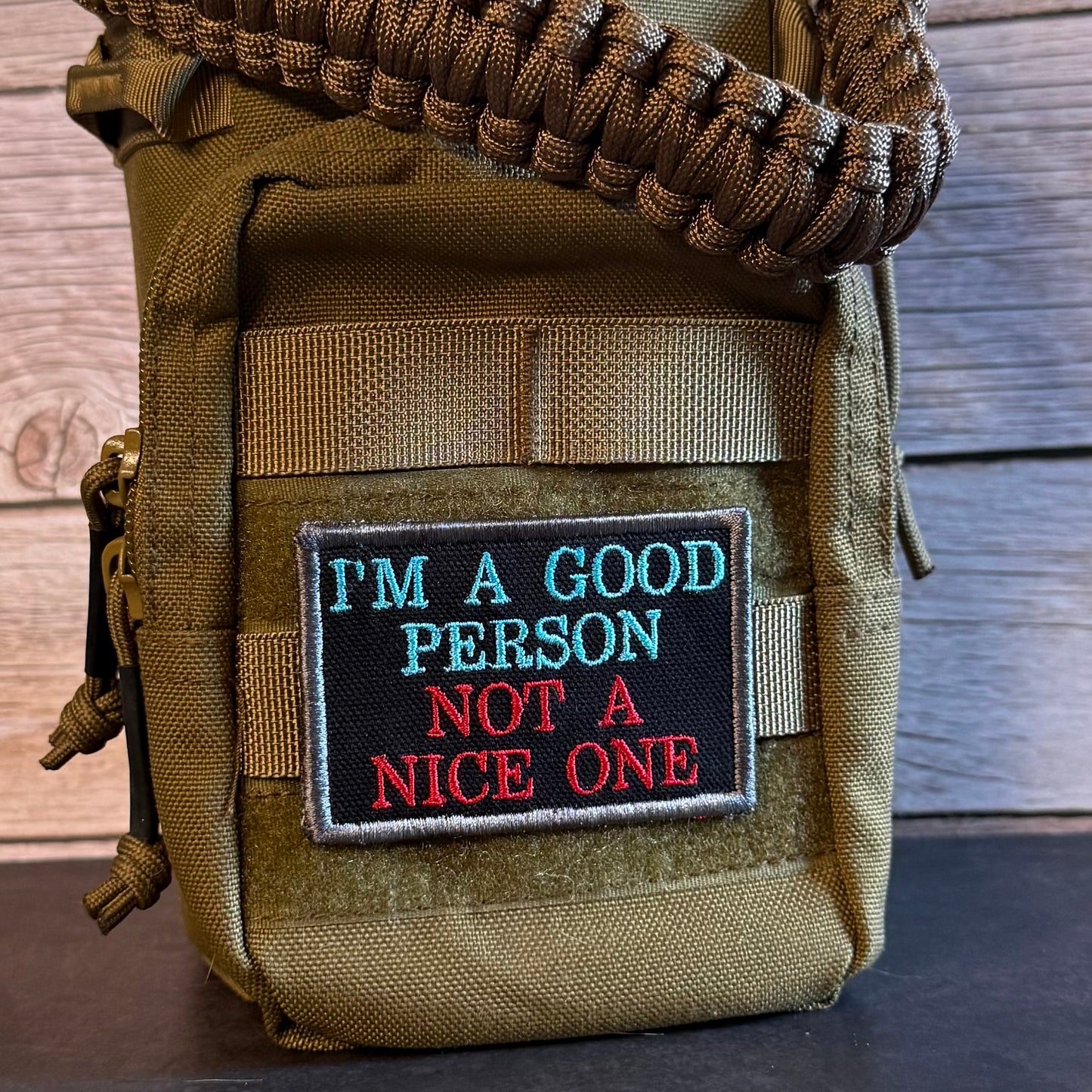 A tactical pouch in olive drab/khaki nylon with a woven paracord strap and a rectangular embroidered patch that reads 'I'M A GOOD PERSON NOT A NICE ONE' in blue and red on a black background, shown against a wood plank backdrop.