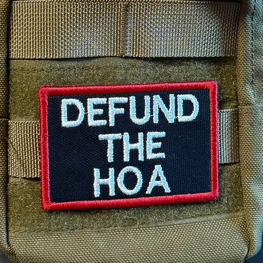 Defund the HOA embroidered morale patch: olive green tactical pouch with a black rectangular patch featuring the white embroidered text 'DEFUND THE HOA' and a red border. A brown paracord strap is attached at the top, and.