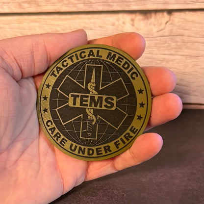 A round tactical medic patch held in a hand, featuring a black and olive green design with the text 'TACTICAL MEDIC CARE UNDER FIRE' and 'TEMS' in the center.