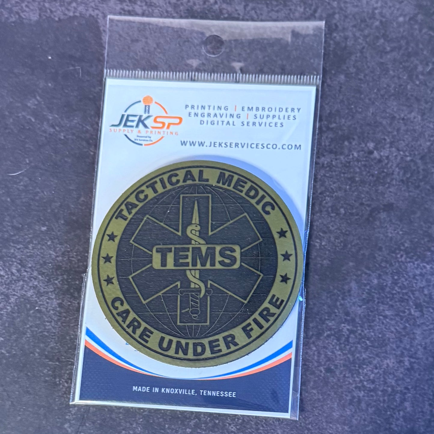 A circular TENS Tactical Medic Care Under Fire patch in olive green and black, mounted on a white backing card with the JEKSP logo, photographed on a dark textured surface.