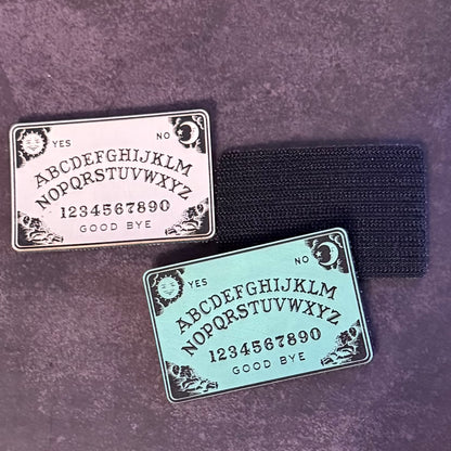 Two rectangular Ouija board style charms with engraved lettering and numbers, placed on a dark textured surface with a black elastic band behind them.