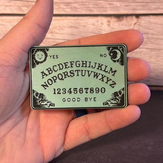 A hand holding a rectangular Ouija board planchette with black engraved symbols and text on a light sage green background, set against a wooden surface.