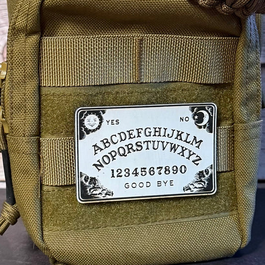 An olive green tactical pouch with a white Ouija board patch on the front, featuring black lettering and symbols, set against a rustic wood background.