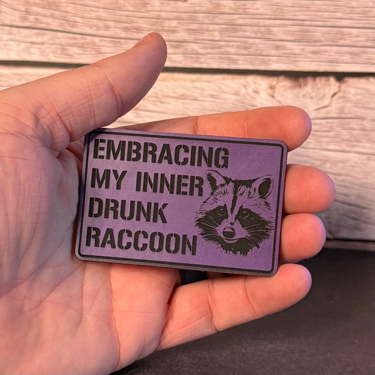 A hand holding a rectangular purple patch with black text reading 'EMBRACING MY INNER DRUNK RACCOON' and a raccoon illustration, against a rustic wood plank background.