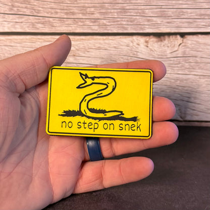 A hand holding a yellow rectangular sign with a black snake illustration and the text 'no step on snek' against a rustic wood plank background.