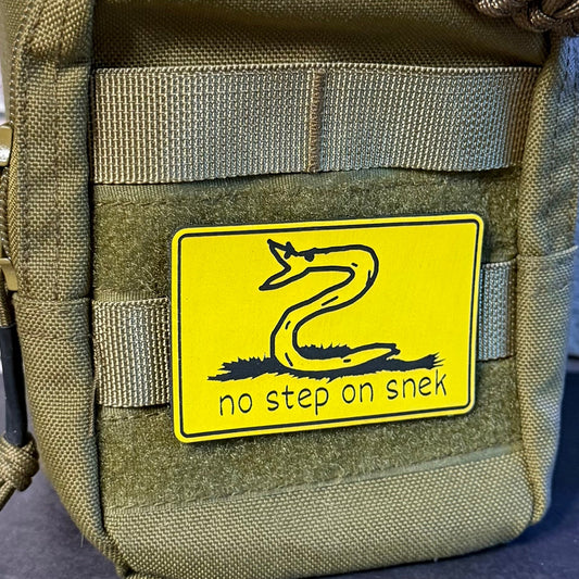 A close-up of an olive green tactical pouch featuring a bright yellow no step on snek rectangular patch with a black snake graphic and the text 'no step on sneak'.