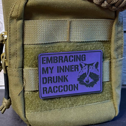 A close-up of a tactical pouch in olive drab green with a purple rectangular patch that reads 'EMBRACING MY INNER DRUNK RACCOON' alongside a raccoon illustration. A brown paracord strap is threaded through the top zipper.