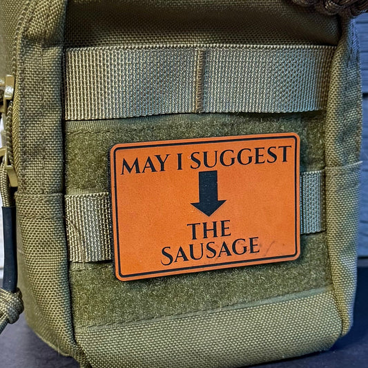 An olive green tactical pouch with a brown faux-leather patch that reads 'MAY I SUGGEST THE SAUSAGE' with a black arrow, shown against a dark wood background.