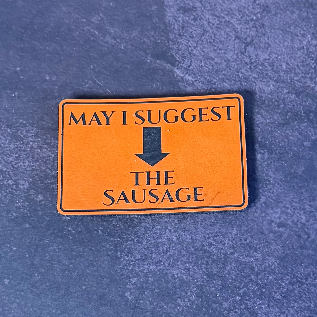 An orange rectangular patch with black text reading 'MAY I SUGGEST' above a downward arrow and 'THE SAUSAGE' below, placed on a textured blue surface.