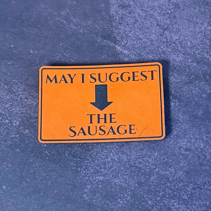 An orange rectangular patch with black text reading 'MAY I SUGGEST' above a downward arrow and 'THE SAUSAGE' below, placed on a textured blue surface.