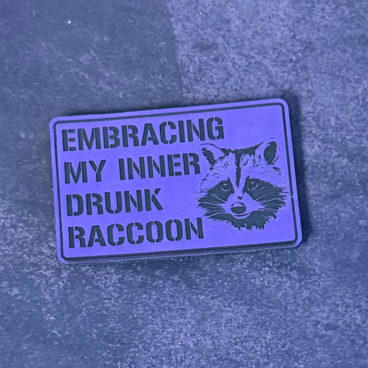 A rectangular enamel pin with a purple background and black text reading 'EMBRACING MY INNER DRUNK RACCOON' alongside a black raccoon illustration, resting on a textured gray surface.