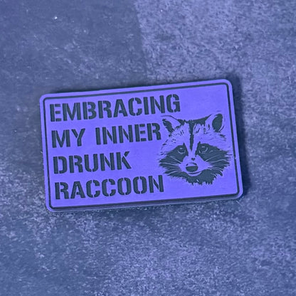 A rectangular enamel pin with a purple background and black text reading 'EMBRACING MY INNER DRUNK RACCOON' alongside a black raccoon illustration, resting on a textured gray surface.