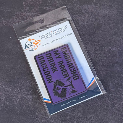 A purple rectangular embroidered patch with black text reading 'EMBRACING MY INNER DRUNK RACCOON' and a black raccoon illustration, sealed in a clear plastic package with a white backing card branded JEK Service Co., photographed on a dark textured surface.
