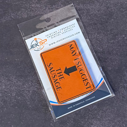 An orange rectangular novelty sign with black text reading 'MAY I SUGGEST THE SAUSAGE' and a black arrow, sealed in clear retail packaging with a visible JKS Service Co. logo on the backing card, photographed on a dark textured surface.