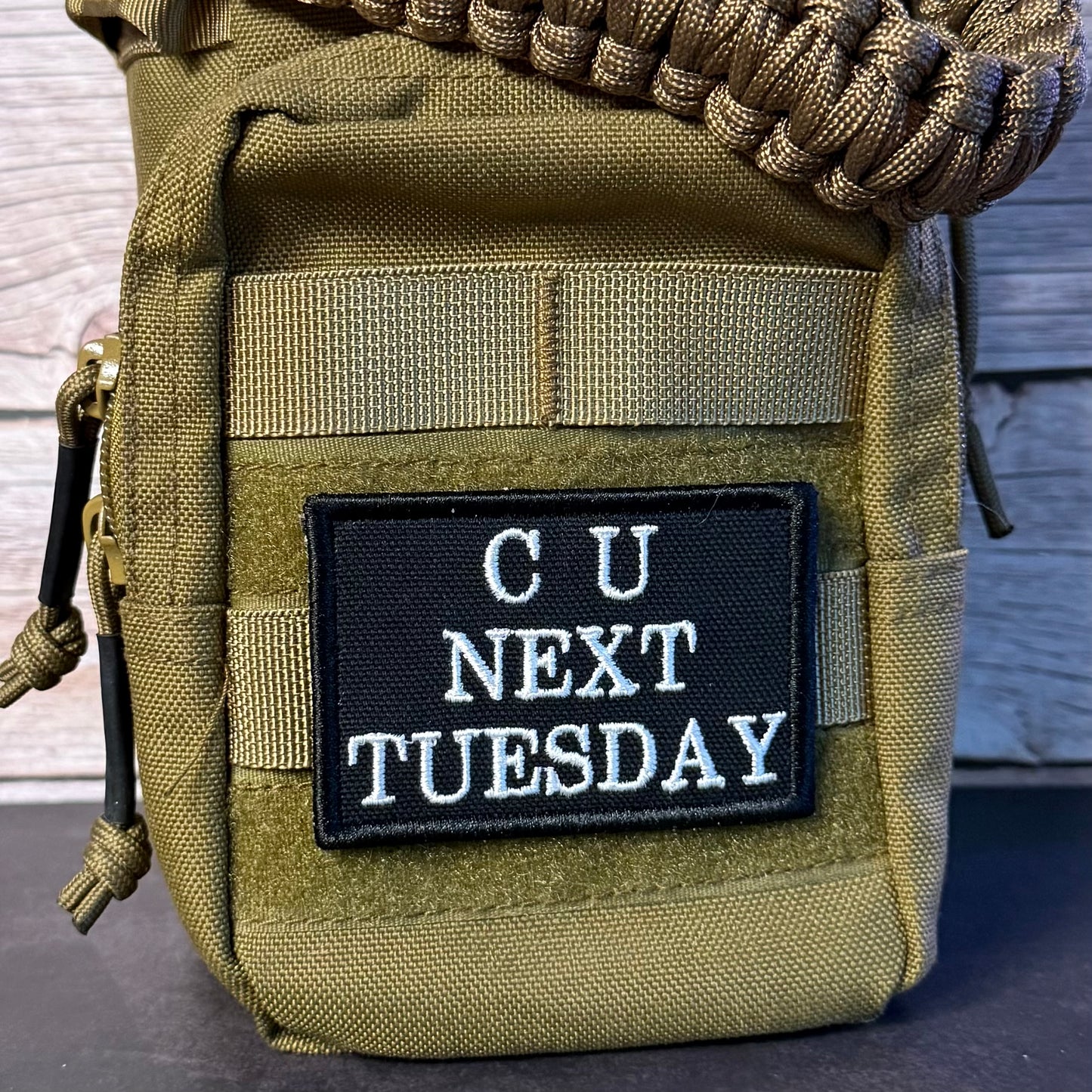 An olive drab tactical pouch with a black rectangular patch that reads 'CU NEXT TUESDAY' in white text, shown close up against a rustic wood background.