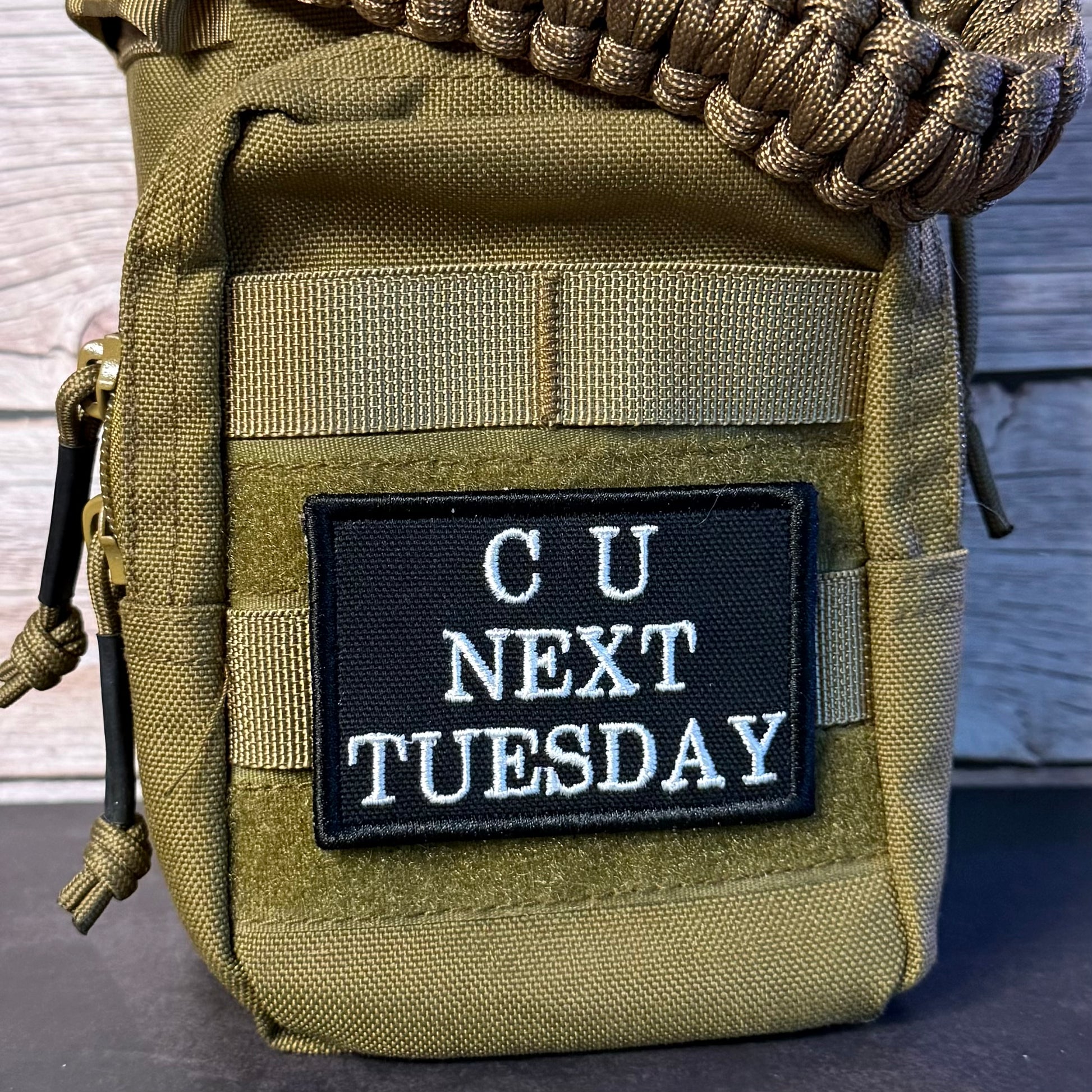 An olive drab tactical pouch with a black rectangular patch that reads 'CU NEXT TUESDAY' in white text, shown close up against a rustic wood background.