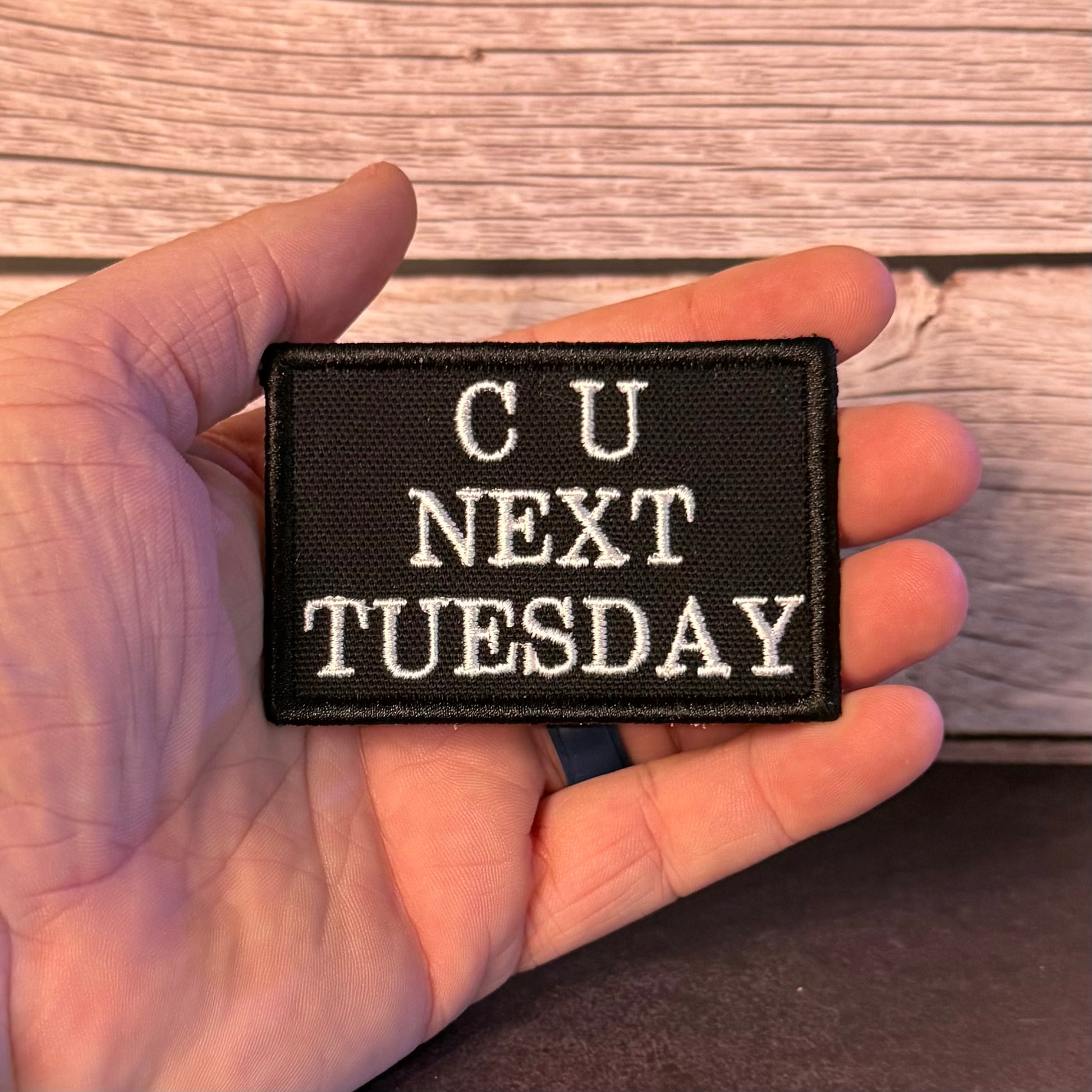 A hand holding a small black embroidered patch with white text that reads 'CU NEXT TUESDAY' against a rustic wood plank background.