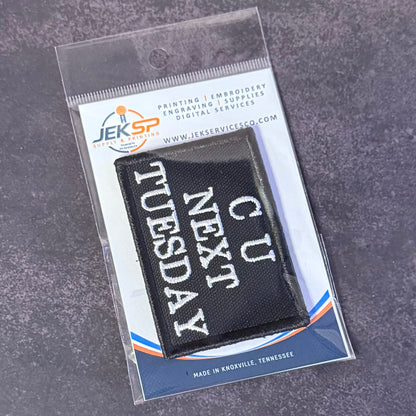 A black rectangular embroidered patch with white text reading 'CU NEXT TUESDAY' inside a clear plastic retail package on a gray textured background. The package card shows the JEKSP brand and website.