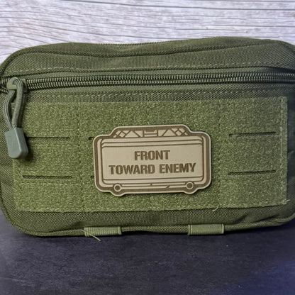 An olive green tactical pouch with a top zipper and a tan rubber patch that reads 'FRONT TOWARD ENEMY' in black text, shown on a dark surface.