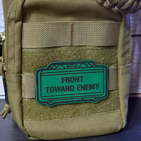 A close-up of a tactical pouch in olive drab and tan webbing, featuring a green rubber patch with the text 'FRONT TOWARD ENEMY' in black.