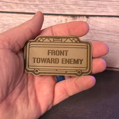 A hand holding a bronze-colored metal key tag with the engraved text 'FRONT TOWARD ENEMY' against a rustic wooden background.