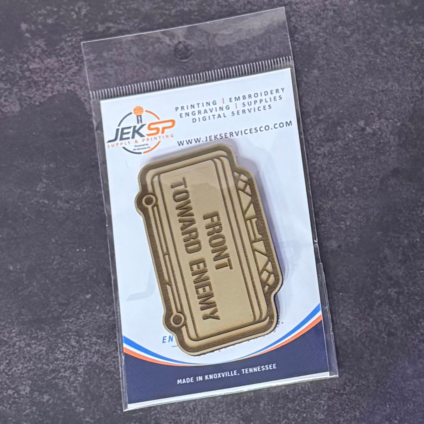 A gold-toned metal lapel pin shaped like a ticket, printed with the words 'FRONT TOWARD ENEMY,' displayed in its original clear plastic packaging on a dark textured background. The packaging card features the Jeksp Service Supply brand logo.