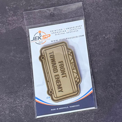 A gold-toned metal lapel pin shaped like a ticket, printed with the words 'FRONT TOWARD ENEMY,' displayed in its original clear plastic packaging on a dark textured background. The packaging card features the Jeksp Service Supply brand logo.