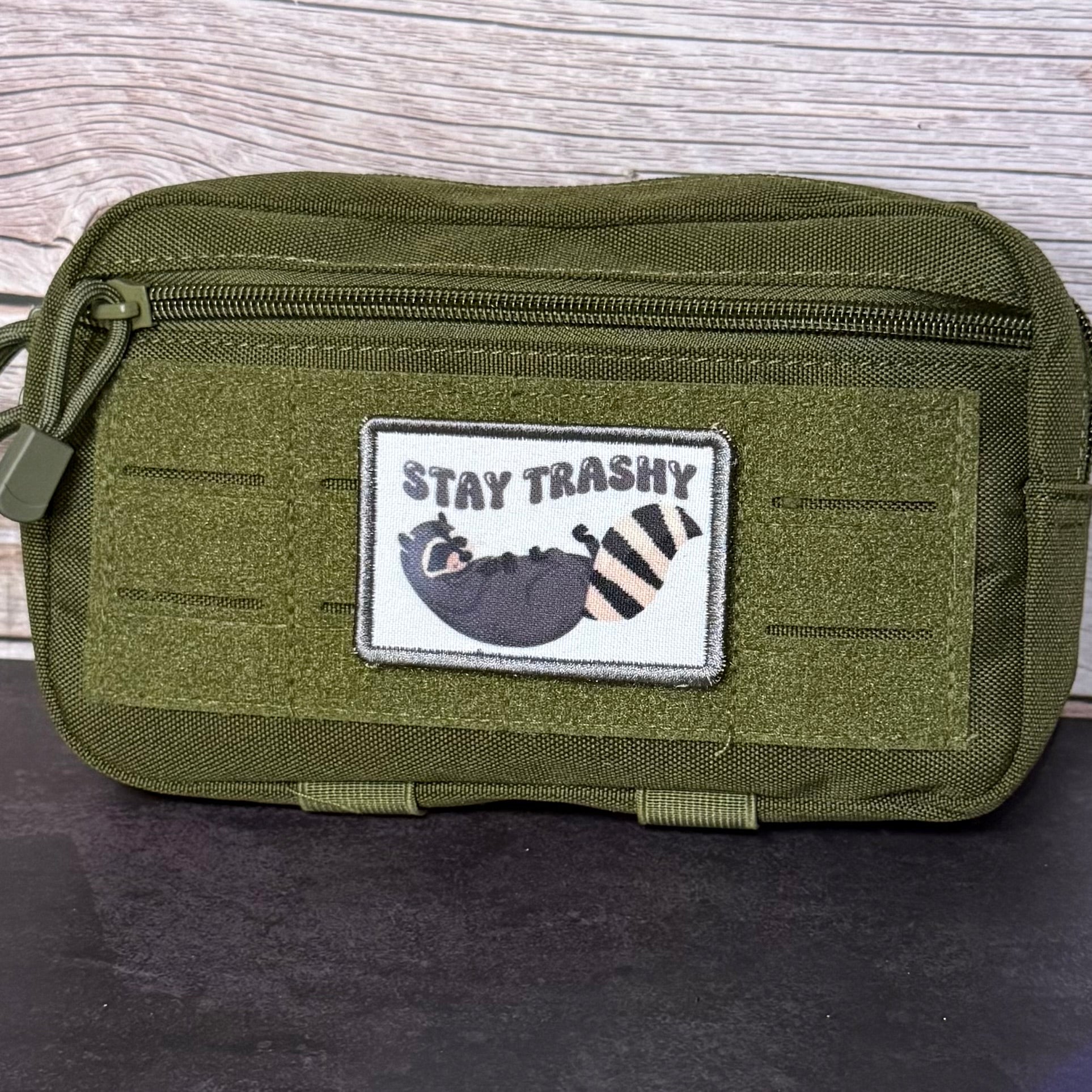 An olive green tactical pouch with a front zipper and a rectangular patch that reads 'STAY TRASHY' above a cartoon raccoon, set against a wood-grain background.