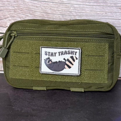 An olive green tactical pouch with a front zipper and a rectangular patch that reads 'STAY TRASHY' above a cartoon raccoon, set against a wood-grain background.