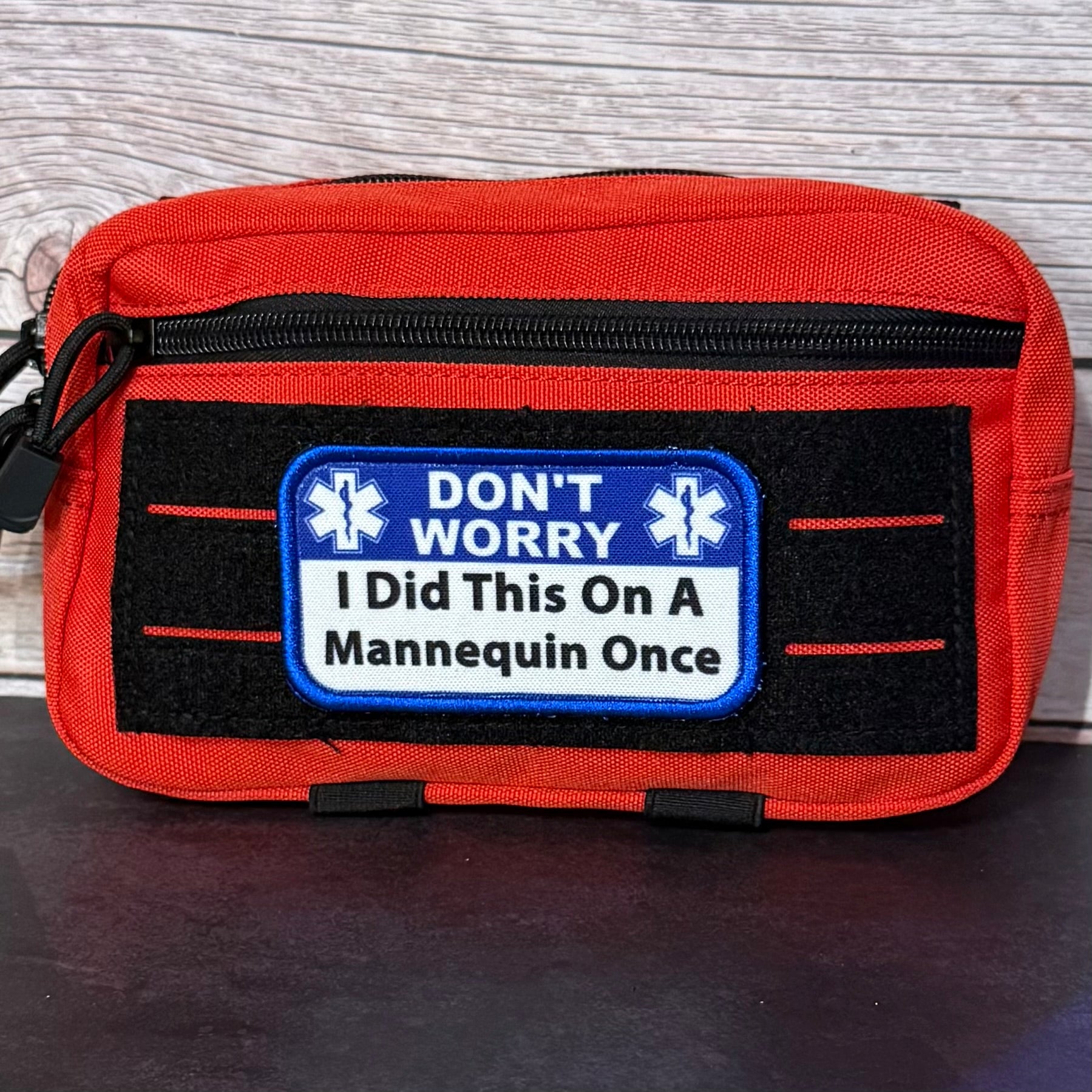 An orange and black first aid pouch with a humorous patch that reads 'DON'T WORRY I Did This On A Mannequin Once' with Star of Life symbols, set against a light wood background.