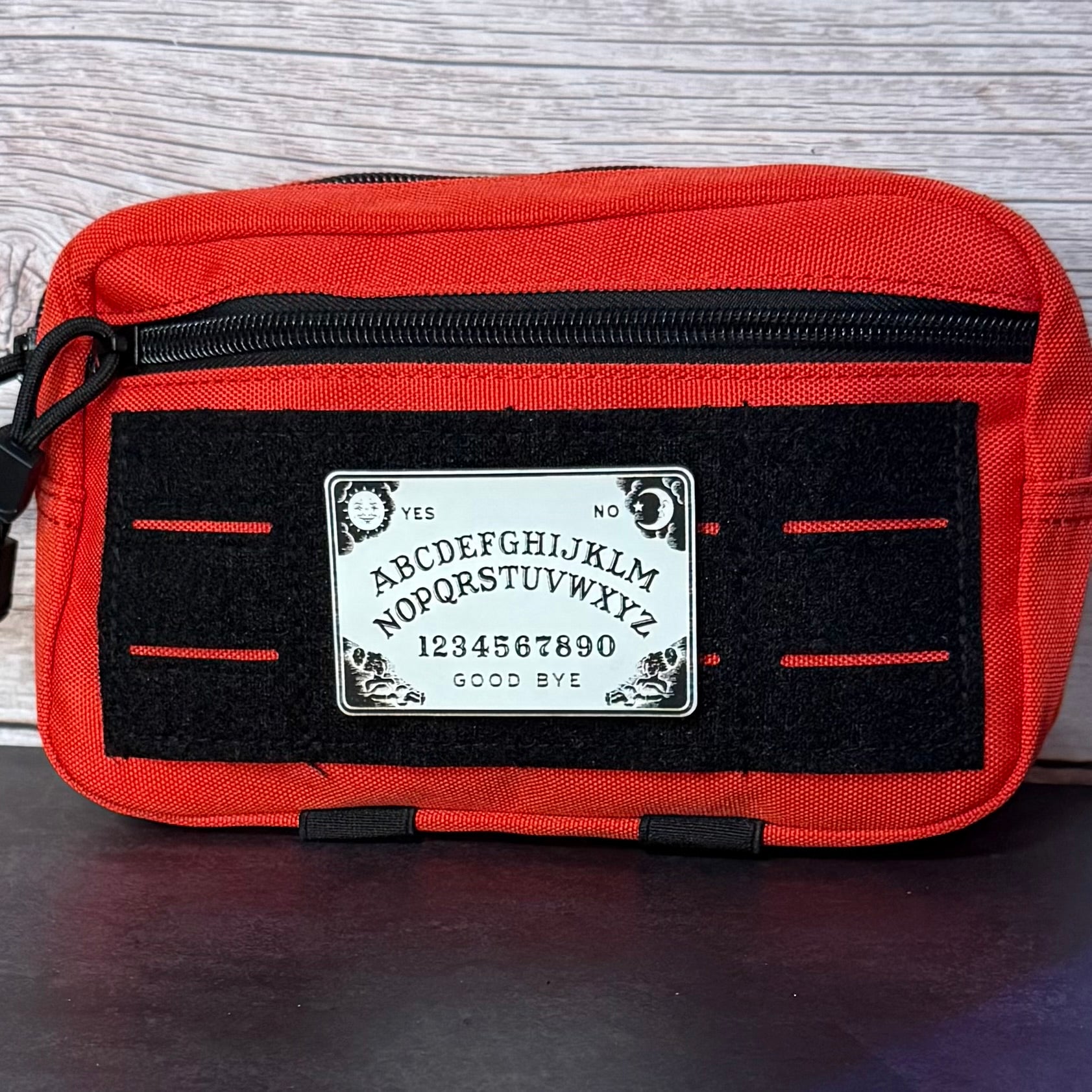 A red canvas pouch featuring a black velcro panel with a white Ouija board graphic in the center, photographed against a light wood background.