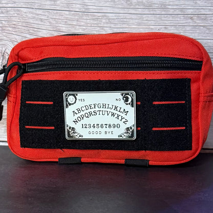 A red canvas pouch featuring a black velcro panel with a white Ouija board graphic in the center, photographed against a light wood background.