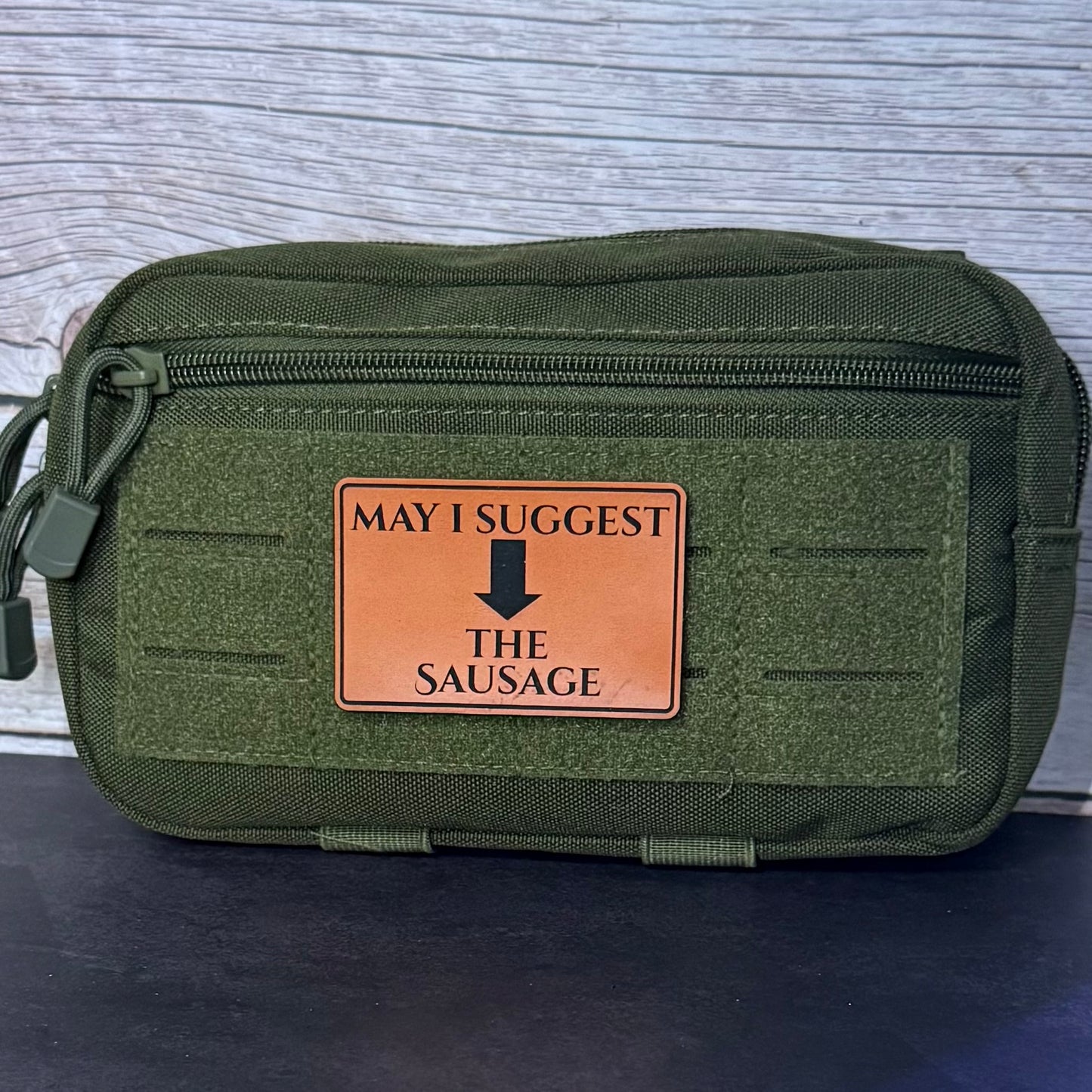 An olive green tactical pouch with a front zipper and a tan leather patch that reads 'MAY I SUGGEST THE SAUSAGE' with a black arrow, set against a gray wood-textured background.
