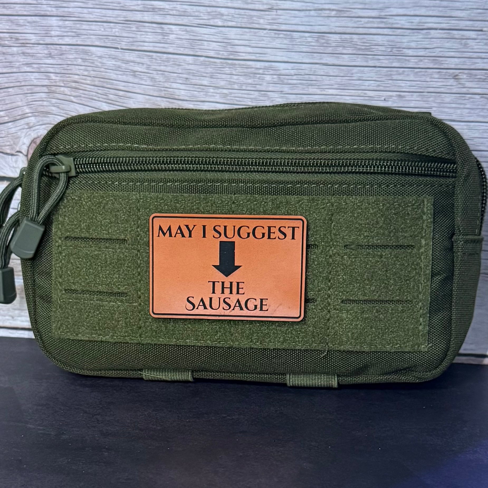 An olive green tactical pouch with a front zipper and a tan leather patch that reads 'MAY I SUGGEST THE SAUSAGE' with a black arrow, set against a gray wood-textured background.