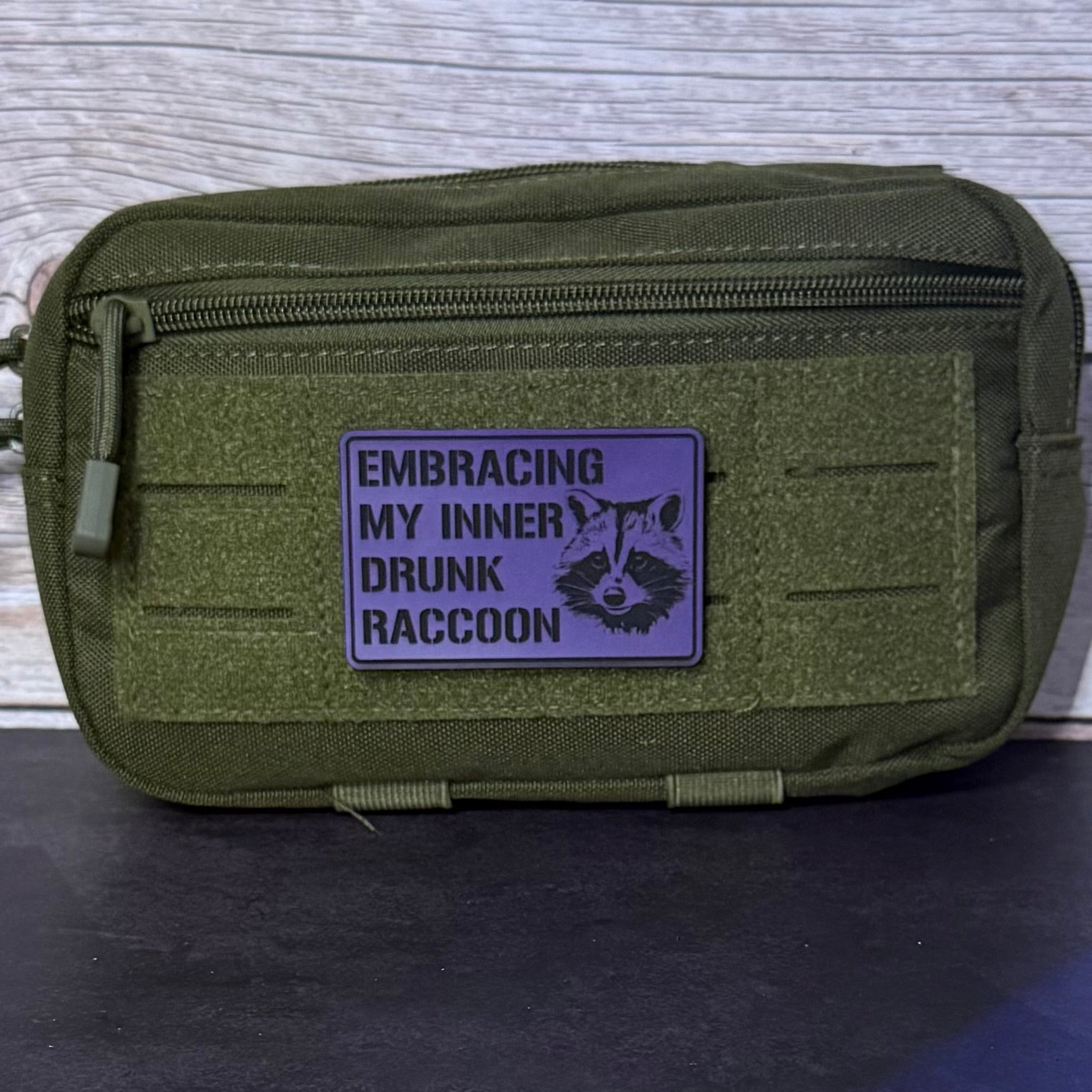 An olive green tactical pouch with a purple rectangular patch that reads 'EMBRACING MY INNER DRUNK RACCOON' with a raccoon graphic, shown against a gray wood-textured background.