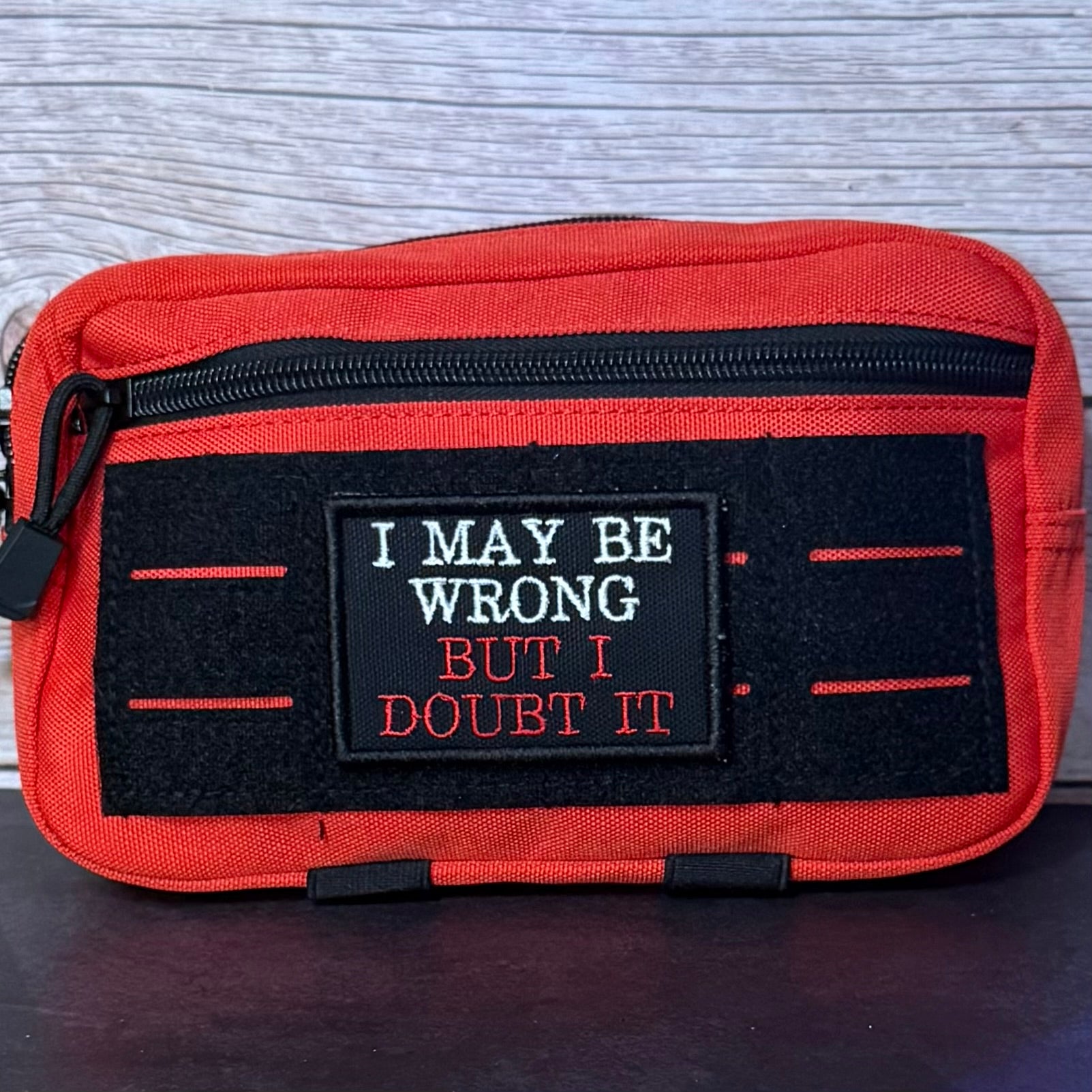 An red canvas pouch with a black zipper and a black velcro patch that reads 'I MAY BE WRONG BUT I DOUBT IT' in white and orange text, shown against a gray wood background.
