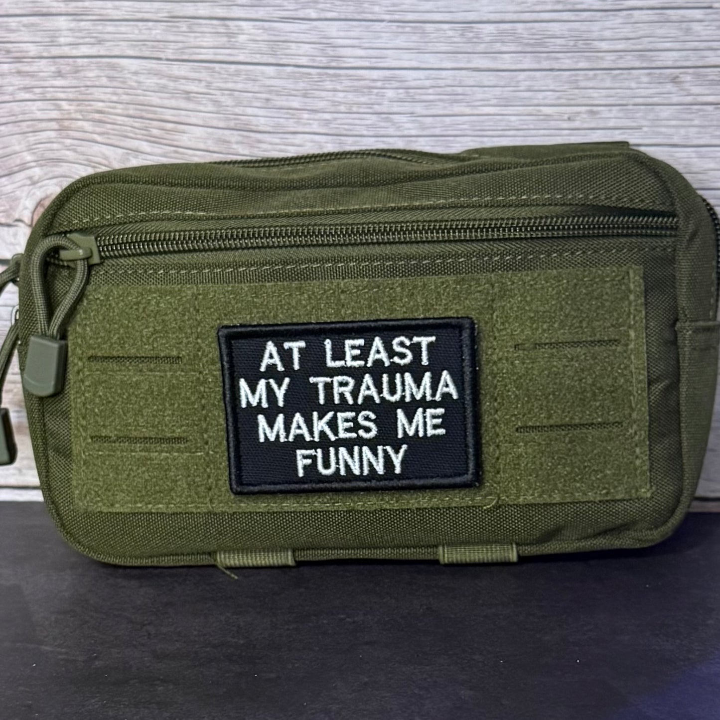 An olive green tactical pouch with a black rectangular patch featuring white embroidered text that reads 'AT LEAST MY TRAUMA MAKES ME FUNNY'.