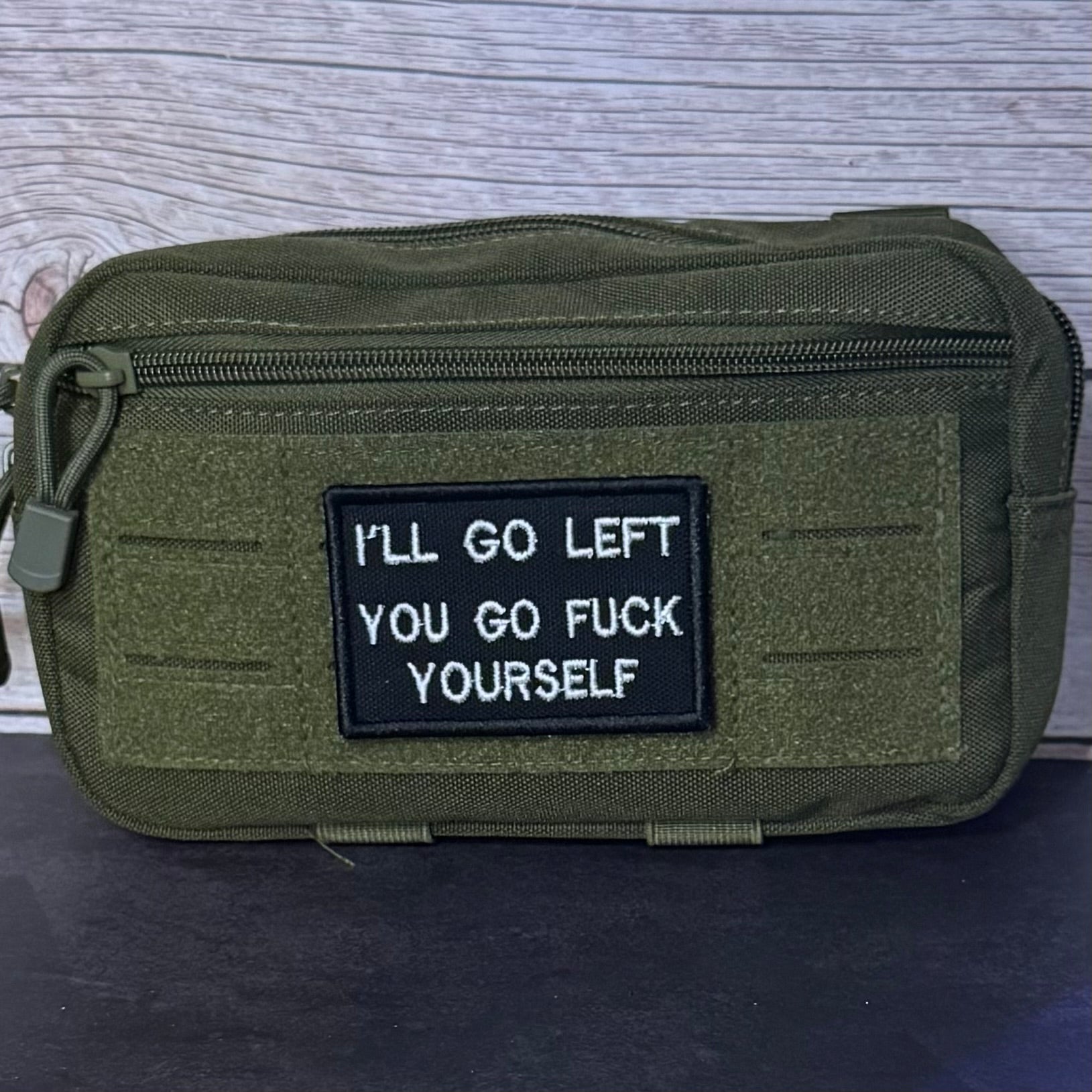 An olive green tactical pouch with a black rectangular patch featuring the white text 'I'LL GO LEFT YOU GO FUCK YOURSELF', set against a light gray wood background.