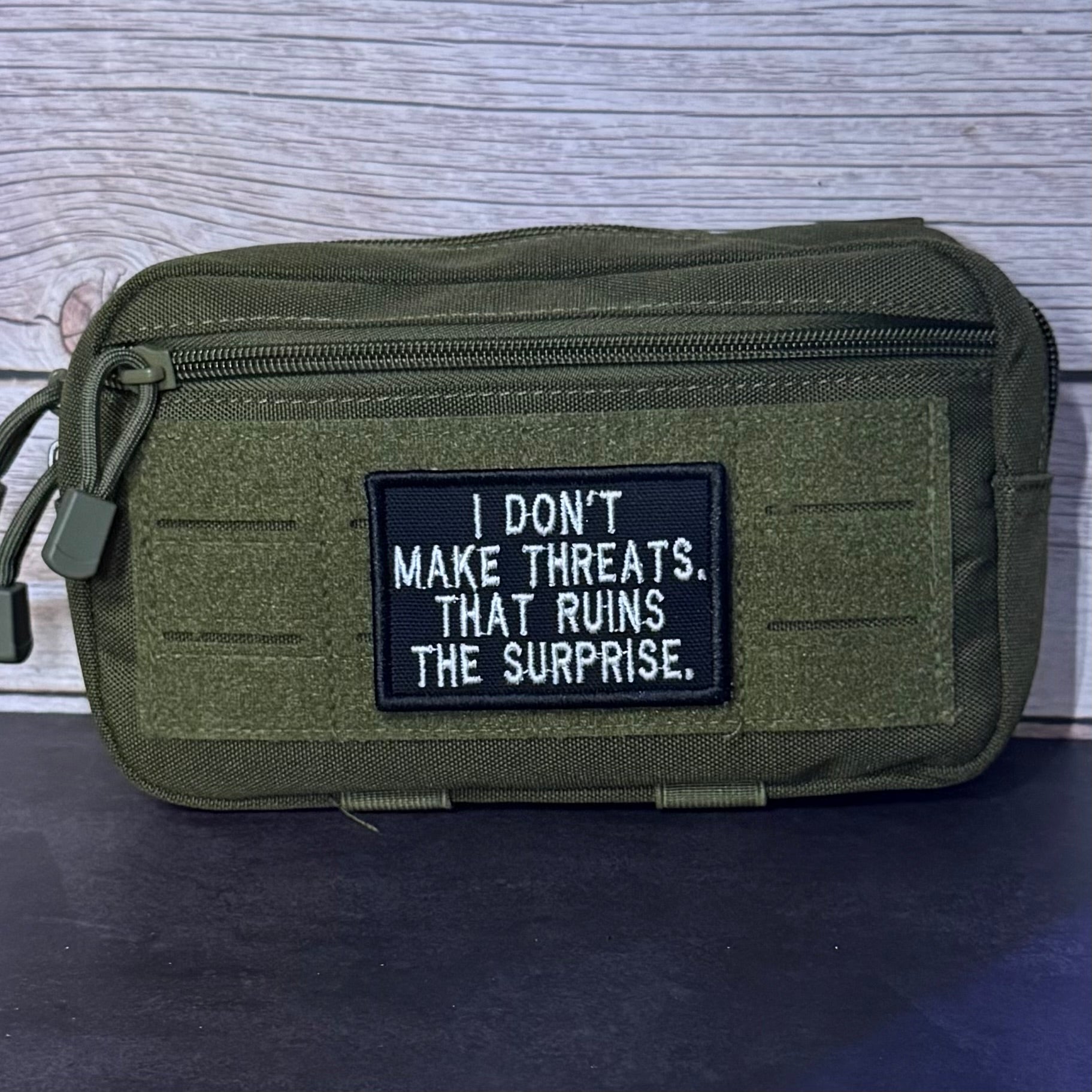An olive green tactical pouch with a black rectangular patch featuring white embroidered text that reads, 'I DON'T MAKE THREATS. THAT RUINS THE SURPRISE.