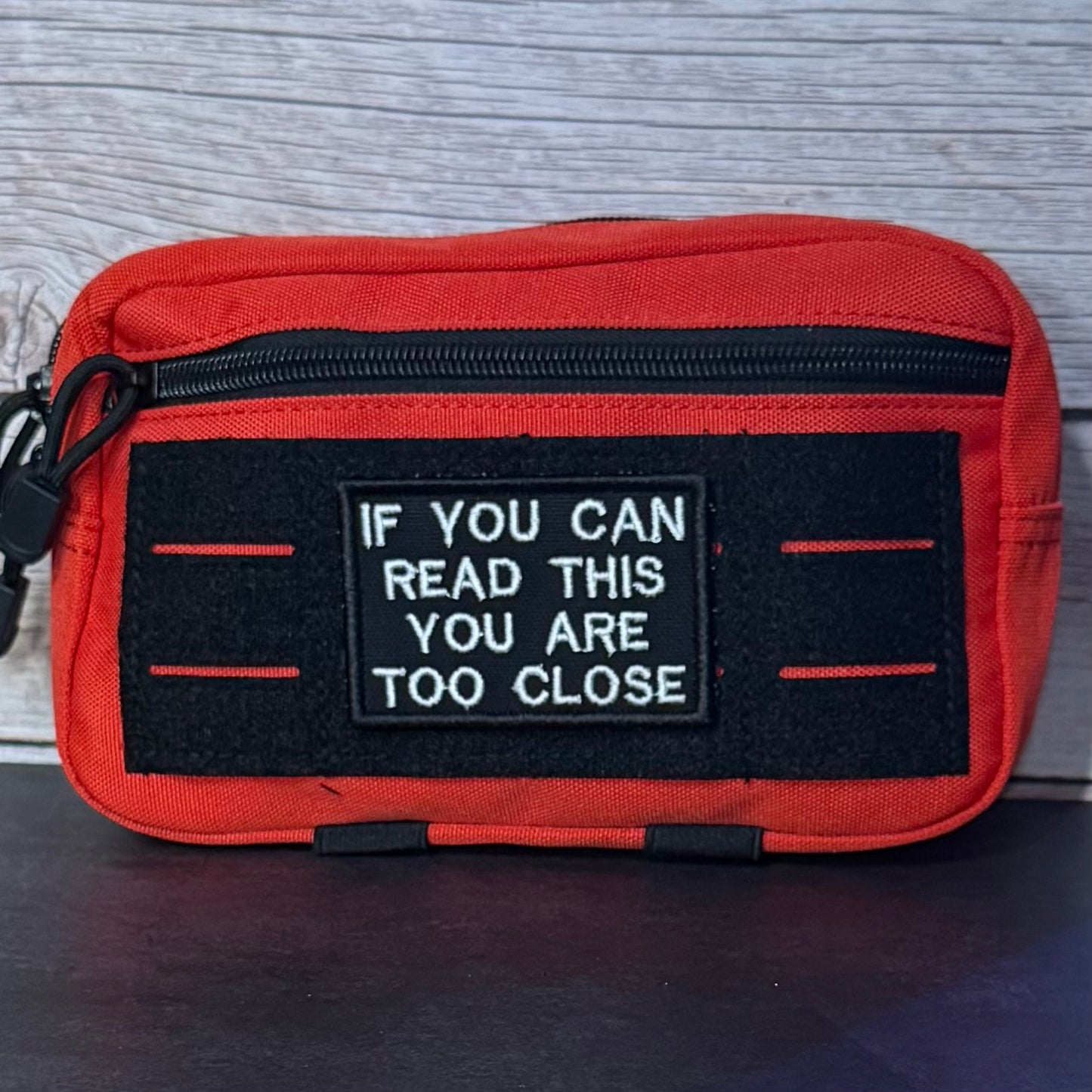 A red and black zippered pouch with a black hook-and-loop patch in the center that reads 'IF YOU CAN READ THIS YOU ARE TOO CLOSE' in white text, shown against a gray wood background.