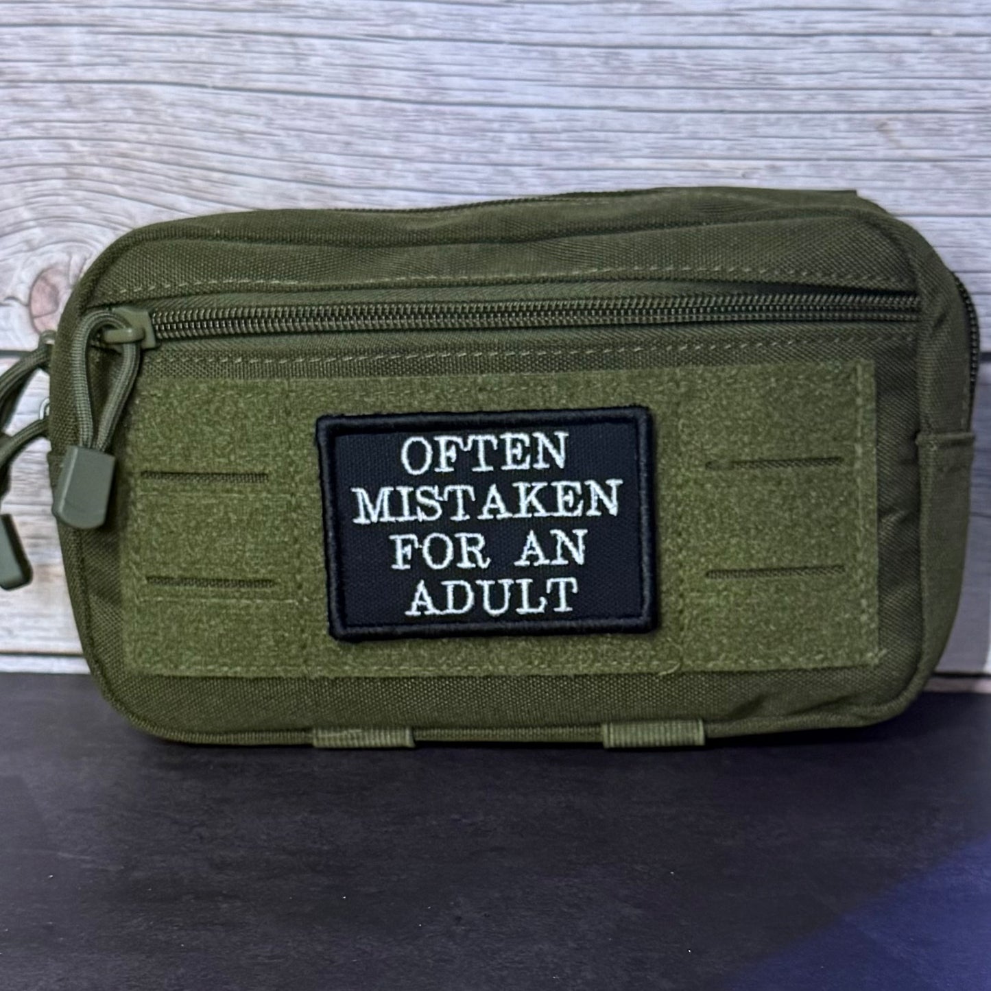An olive green tactical pouch with a black rectangular patch that reads 'OFTEN MISTAKEN FOR AN ADULT' in white text, shown against a light wood background.