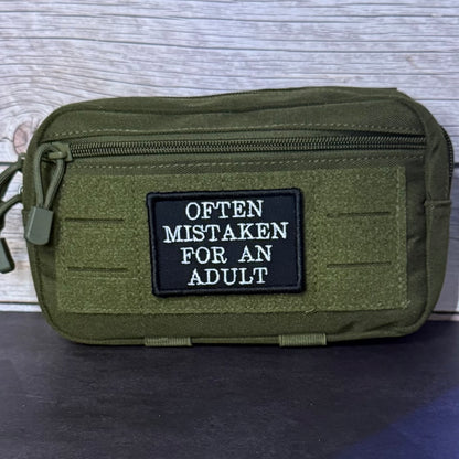 An olive green tactical pouch with a black rectangular patch that reads 'OFTEN MISTAKEN FOR AN ADULT' in white text, shown against a light wood background.
