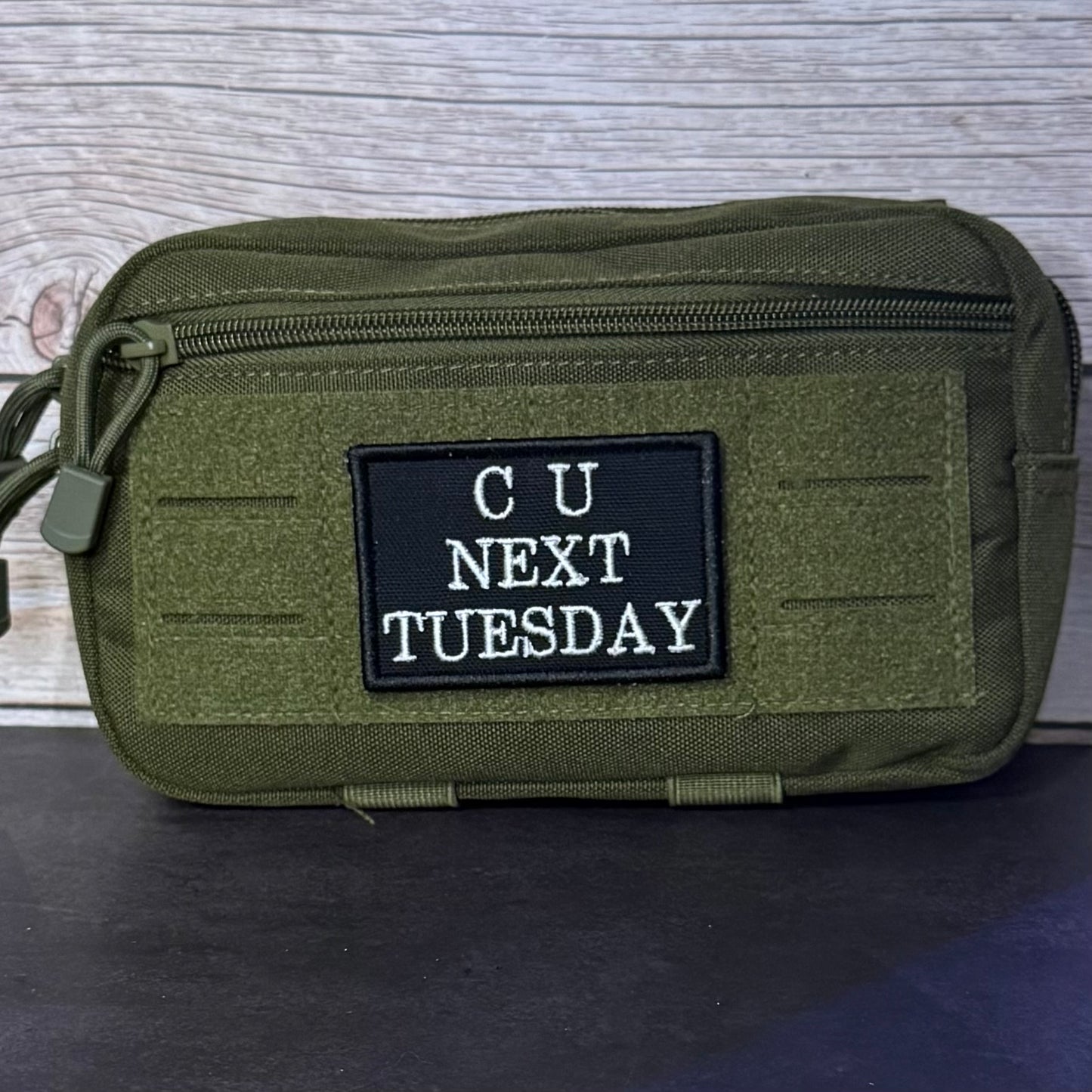 An olive green tactical pouch with a black rectangular patch that reads 'CU NEXT TUESDAY' in white text, shown against a gray wood-grain background.