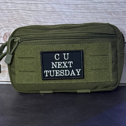 An olive green tactical pouch with a black rectangular patch that reads 'CU NEXT TUESDAY' in white text, shown against a gray wood-grain background.