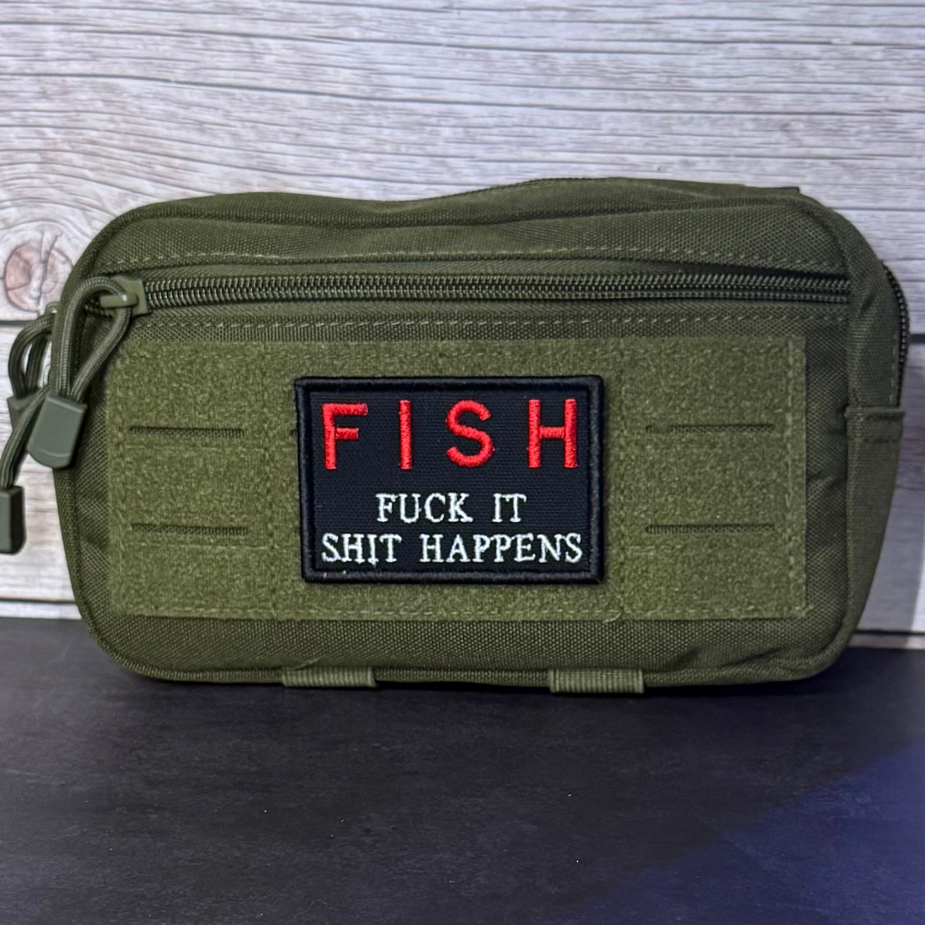 An olive green tactical pouch with a black rectangular patch featuring the word 'FISH' in red and the phrase 'FUCK IT SHIT HAPPENS' in white, set against a light wood background.