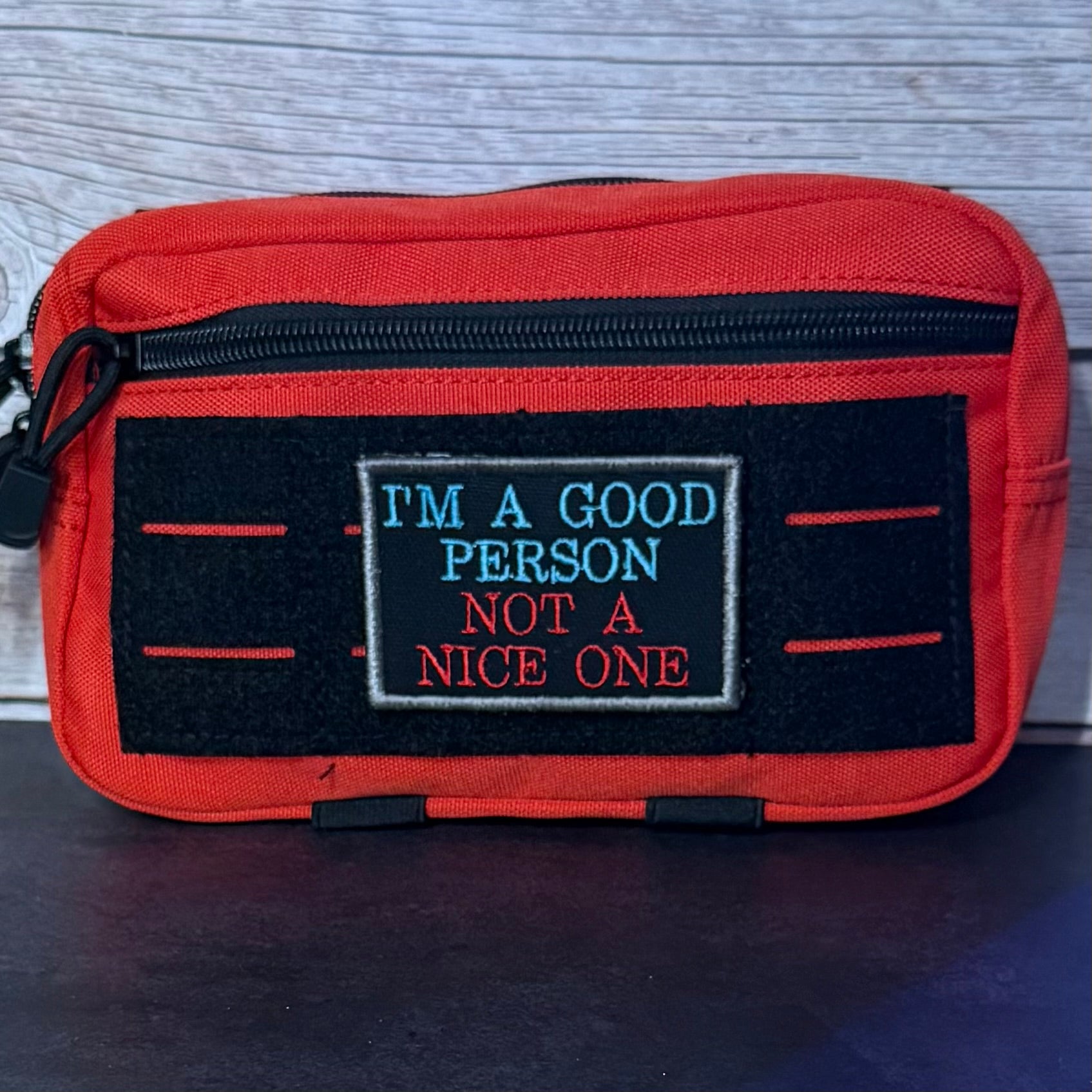 A red canvas pouch with a black front patch that reads 'I'M A GOOD PERSON NOT A NICE ONE' in blue and red embroidery, photographed against a gray wood-textured background.