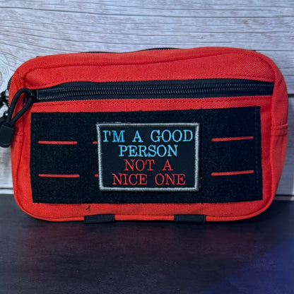 A red canvas pouch with a black front patch that reads 'I'M A GOOD PERSON NOT A NICE ONE' in blue and red embroidery, photographed against a gray wood-textured background.