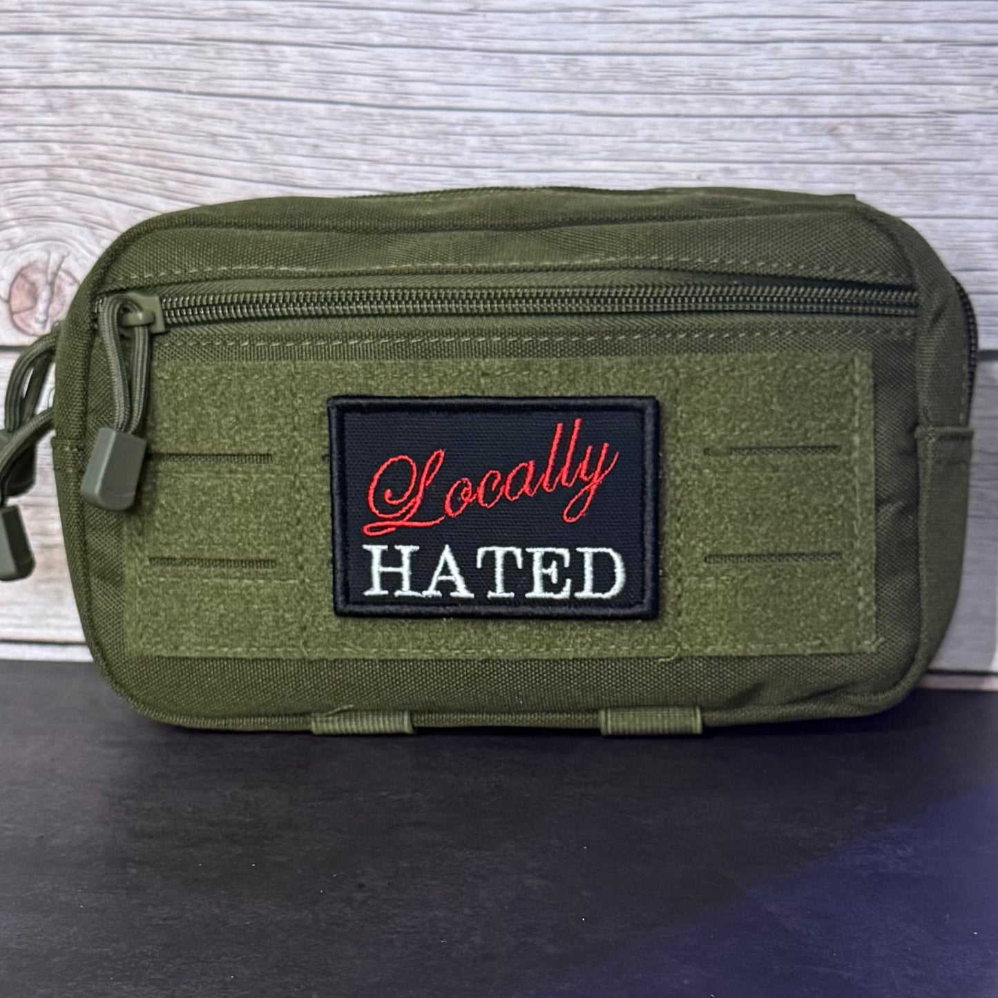 An olive green tactical pouch with a front zippered pocket and a black embroidered patch reading 'Locally HATED' in red and white, set against a light wood background.