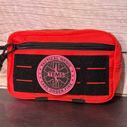 A red tactical first aid pouch with a black velcro panel and a circular pink and black 'Tactical Medic TEMS Care Under Fire' patch, shown against a wood-grain background.